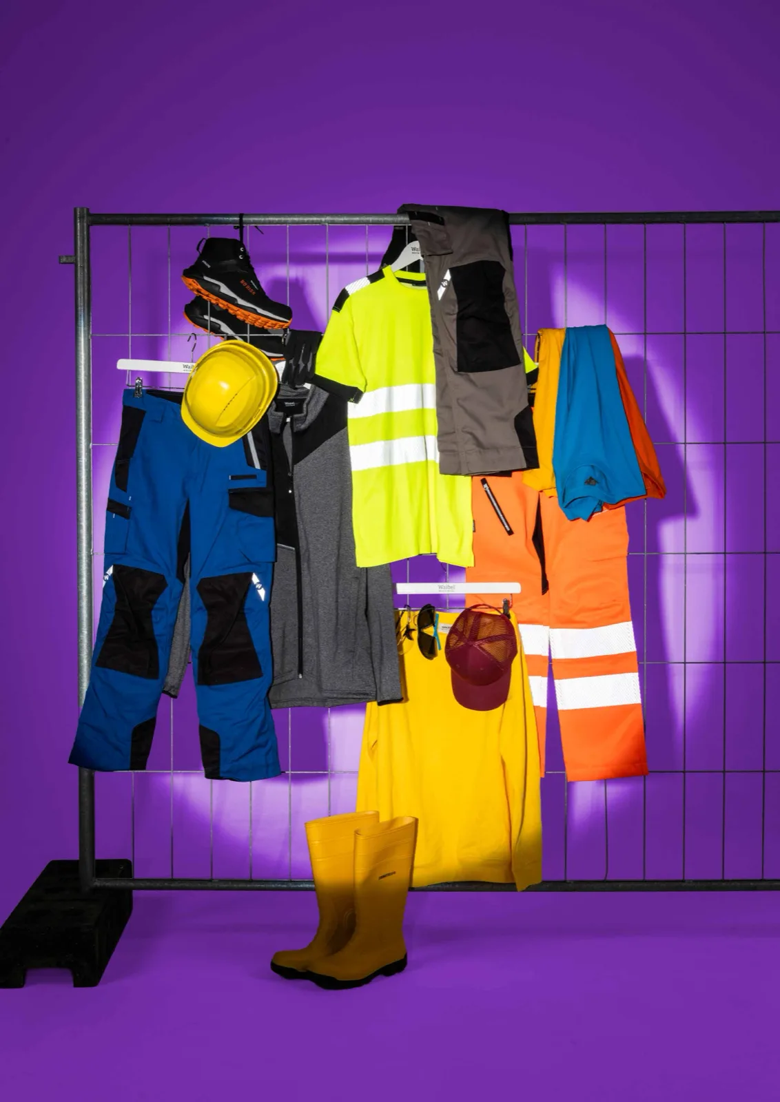 Workwear-Sortiment an einem Bauzaun: Arbeitshosen, Warnschutzkleidung, Helm und Stiefel